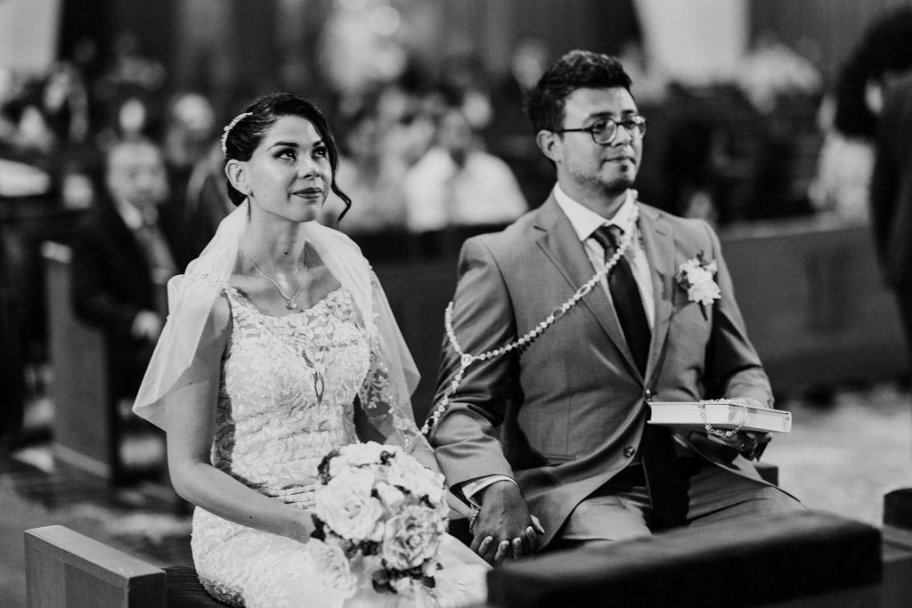 Black and white photo of bride and groom sitting side by side during their Mexico City wedding ceremony, holding hands and draped with a lasso