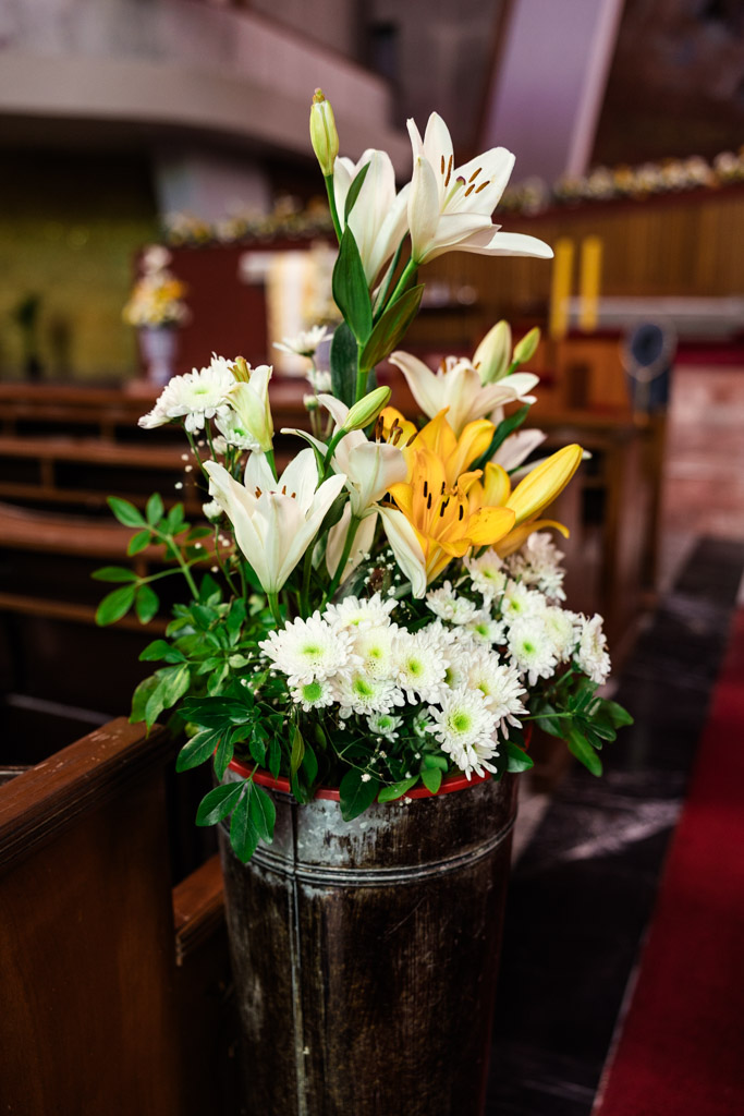 A bouquet of white and yellow flowers with greenery in a rustic vase graces a church