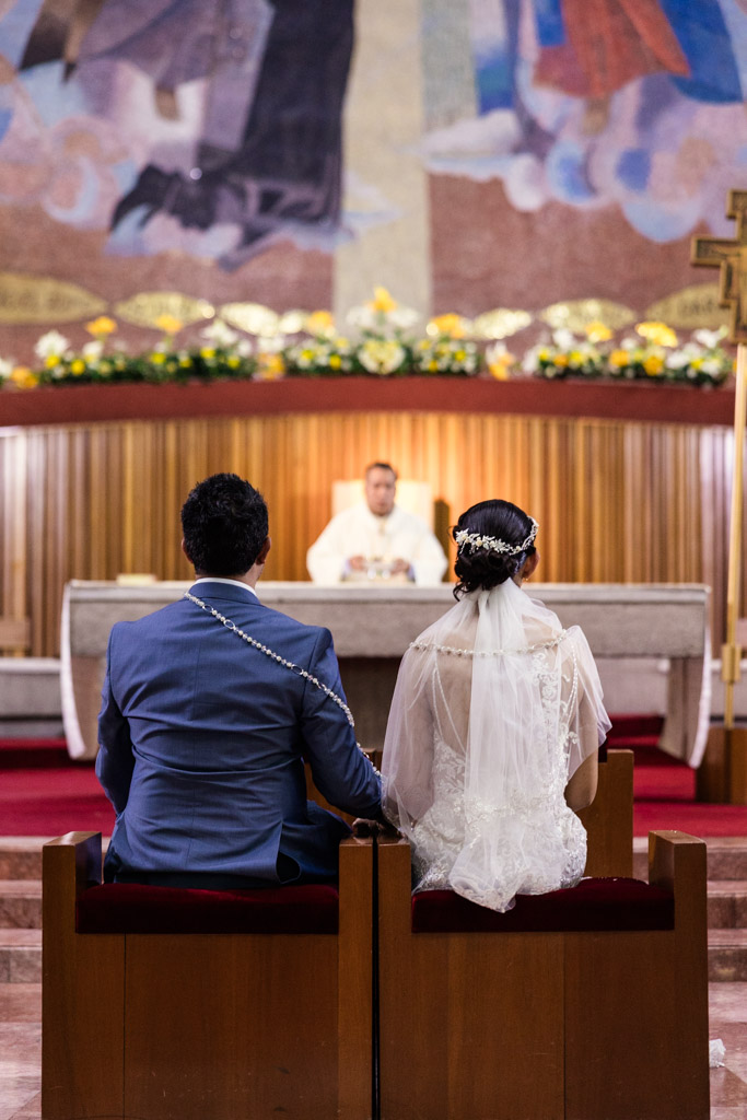A bride and groom sit at the altar facing a priest during their wedding ceremony at Parroquia San Cayetano