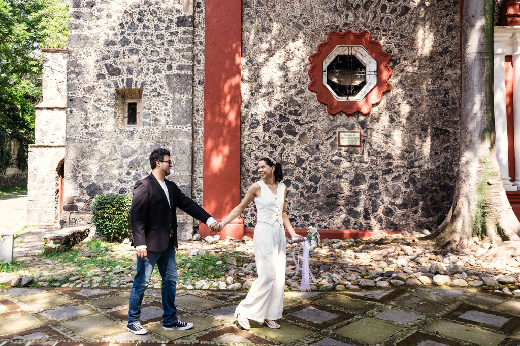 A couple holding hands walks past a stone church with a red-trimmed window, smiling at each other during Mexico City newlywed session