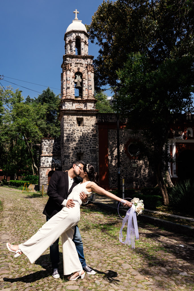 Groom dips his partner in front of a historic stone church in Mexico City's Chimalistac neighborhood on a sunny day