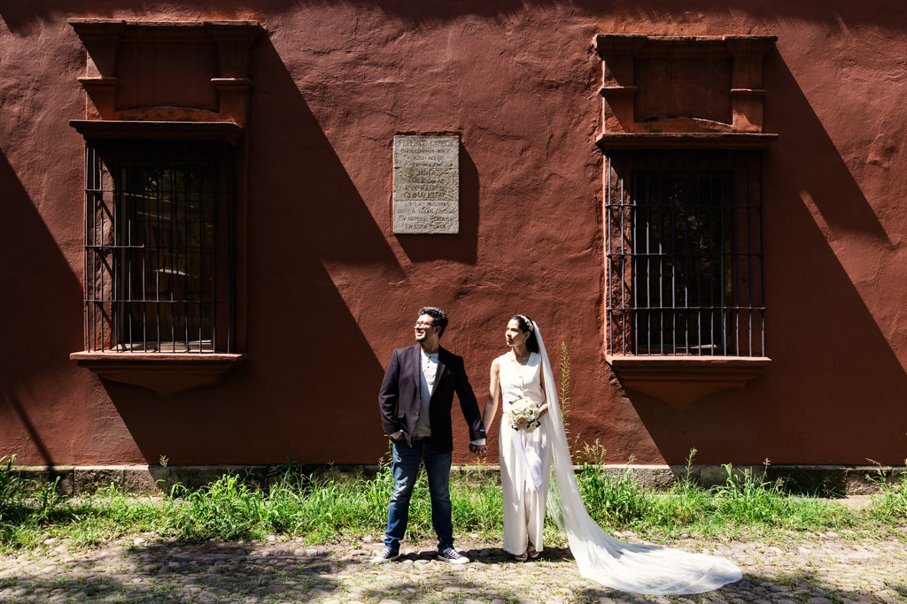 Bride and groom hold hands, bathed in sunlight against a red wall with two windows during Mexico City newlywed session