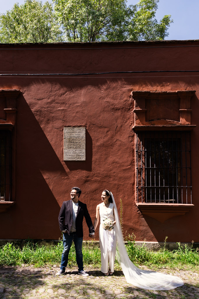 Bride and groom hold hands in front of a red wall with barred windows and a plaque in bright sunlight in Mexico City's Chimalistac neighborhood