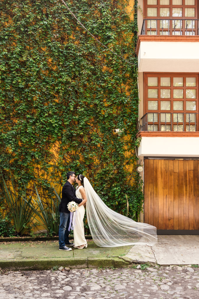 Bride and groom kiss on a sidewalk beside a vine-covered yellow wall and wooden door.in Mexico City's Chimalistac neighborhood