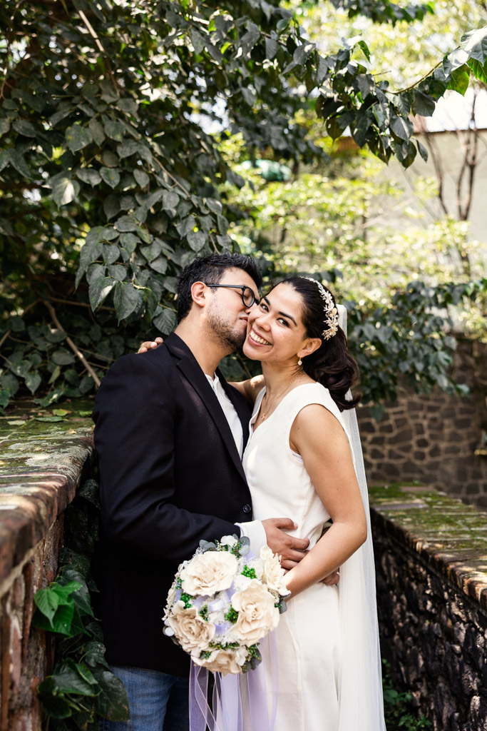 Groom kisses bride's cheek as they stand on a stone bridge in Chimalistac neighborhood during Mexico City newlywed session