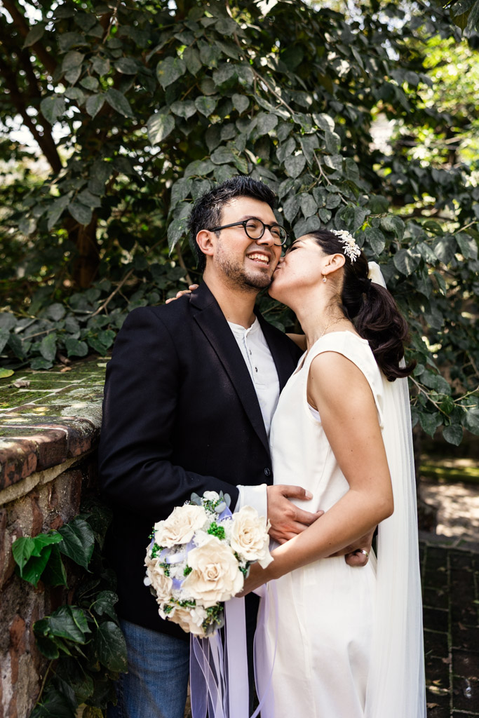 Bride kisses groom’s cheek as they stand outdoors by a brick wall and greenery during Mexico City newlywed session