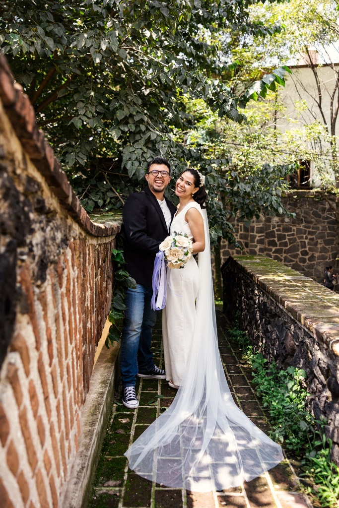 A smiling couple in wedding attire poses on a brick path, surrounded by lush greenery and sunlight during newlwed session in Mexico City's Chimalistac neighborhood