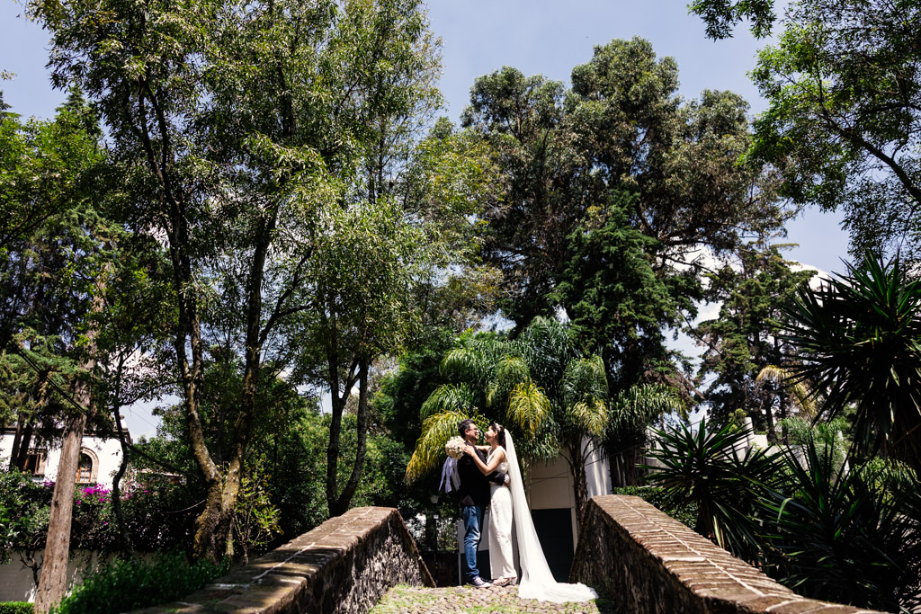 Bride and groom embrace on a stone bridge surrounded by lush green trees under a clear sky during Mexico City newlywed session