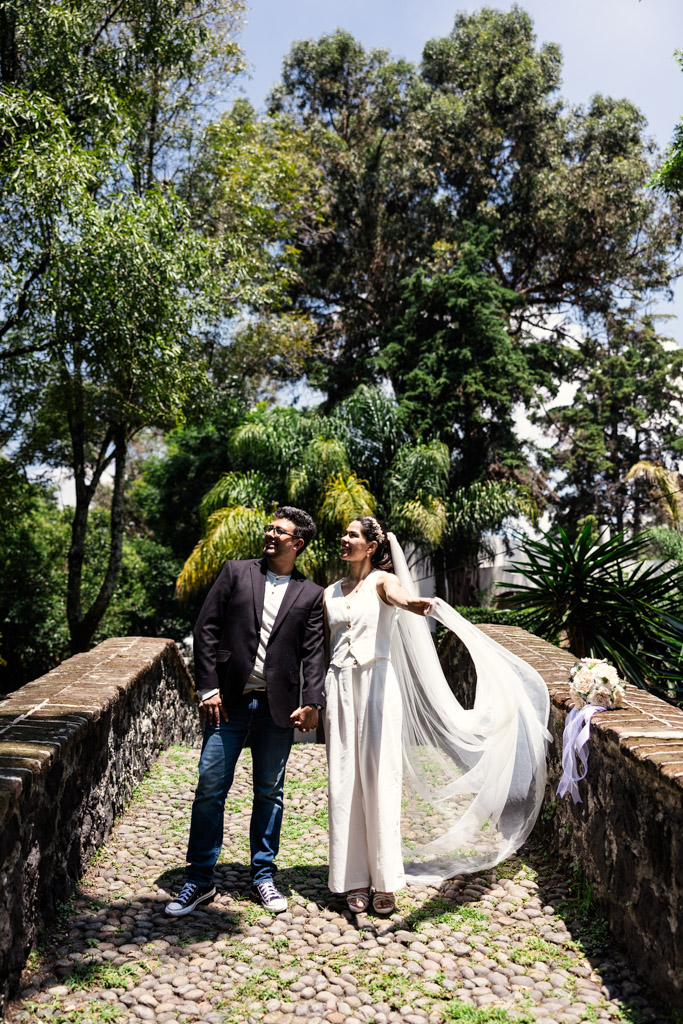 Bride and groom hold hands on a stone bridge, surrounded by lush trees and greenery in Chimalistac neighborhood during Mexico City newlywed session