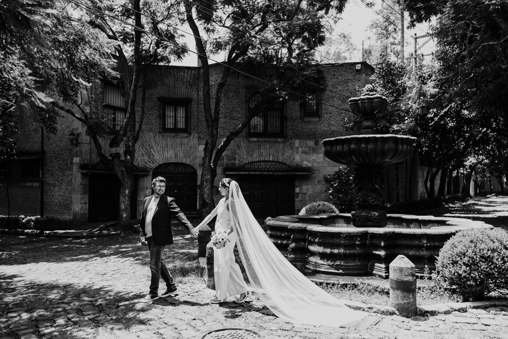 Black and white photo of bride and groom holding hands, walking by a stone fountain on a cobblestone street in Chimalistac neighborhood of Mexico City