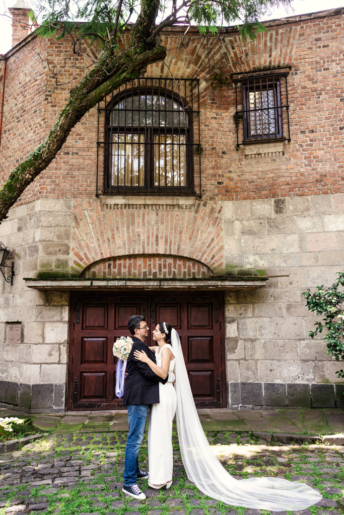 Bride and groom embrace in front of a rustic brick building with arched wooden doors during newlywed session in Chimalistac, Mexico City