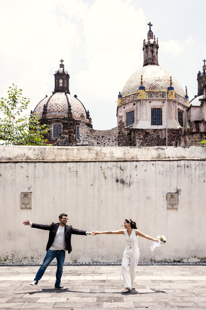 Newlywed couple holds hands, smiling, in front of a white wall and domed church with crosses in the background at Museo de el Carmen
