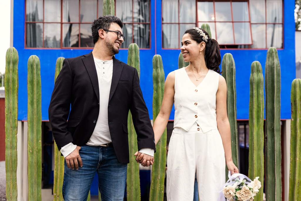 Couple holds hands and smiles in front of tall cacti and a bright blue building at Museo Casa Estudio Diego Rivera y Frida Kahlo in Mexico City