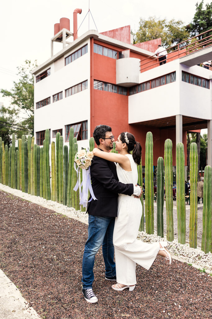 Couple embraces and smiles in front of Museo Casa Estudio Diego Rivera y Frida Kahlo and a row of tall cacti during Mexico City newlywed session