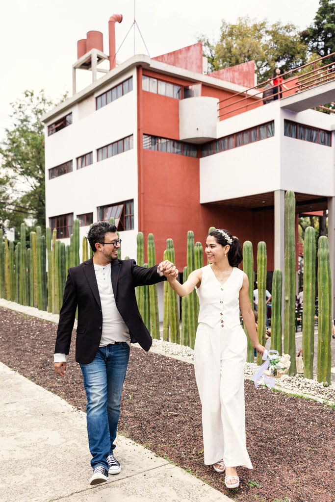 Birde and groom walk hand in hand, smiling, in front of Museo Casa Estudio Diego Rivera y Frida Kahlo during Mexico City newlywed session