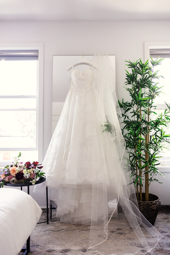 A white wedding dress with a long veil hangs on a mirror in a bright room with bride's bouquet