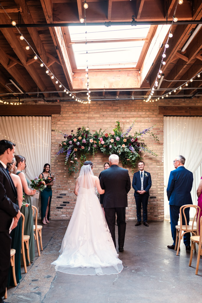 Bride walks down the aisle with her father during her Arbory Chicago wedding ceremony under string lights and a skylight