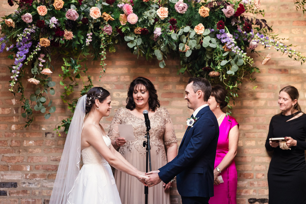 Bride and groom hold hands while bride's mother gives a reading during their wedding ceremony at The Arbory Chicago