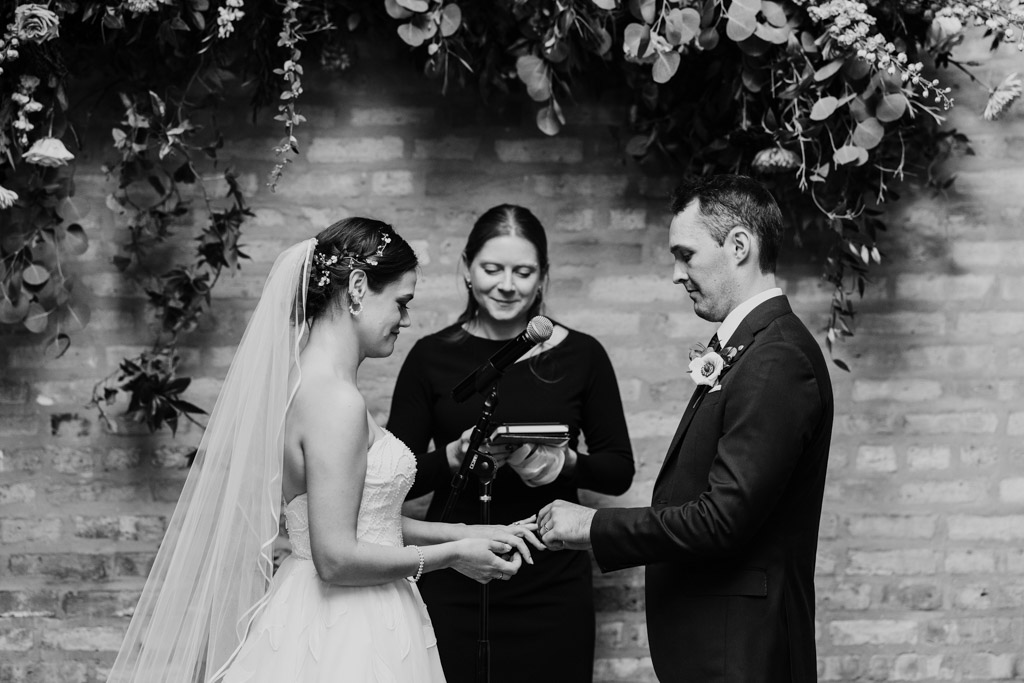 Black and white photo of bride and groom exchanging rings during their wedding ceremony at The Arbory Chicago, with an officiant standing behind them