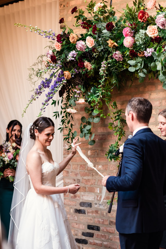 Bride and groom smiling while performing Celtic handfasting during their wedding ceremony at The Arbory Chicago under a floral arch