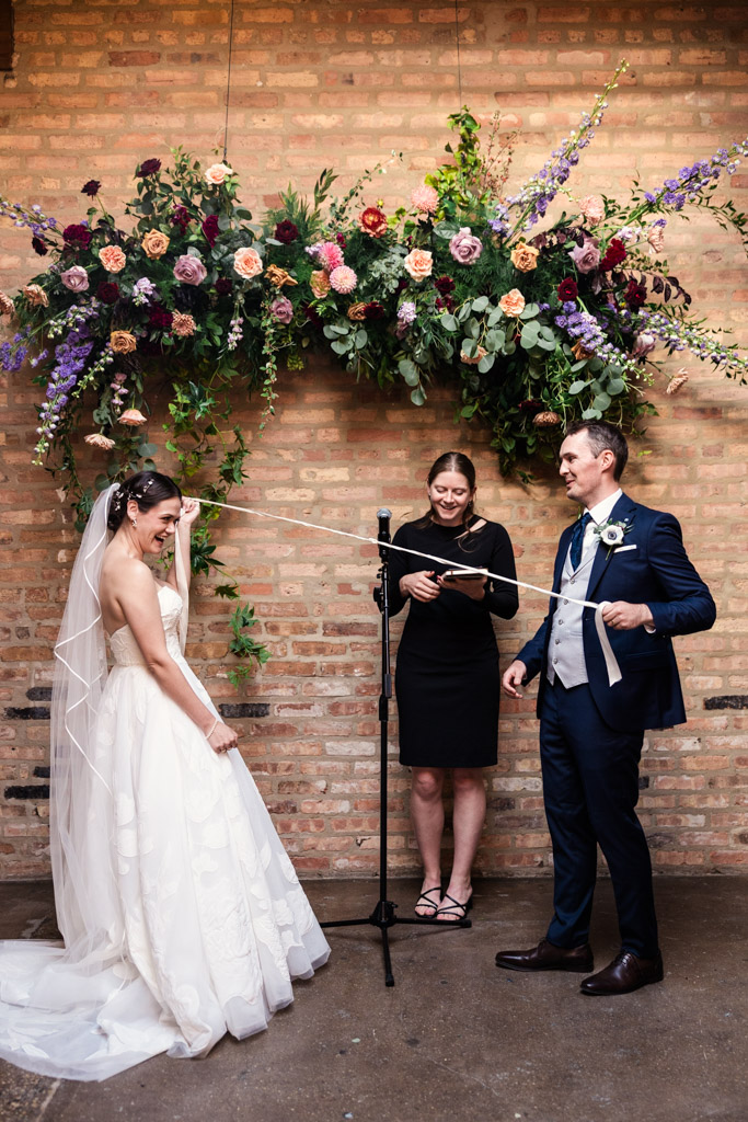 Bride and groom smiling during the handfasting portion of their wedding ceremony, standing under a floral arch at The Arbory Chicago