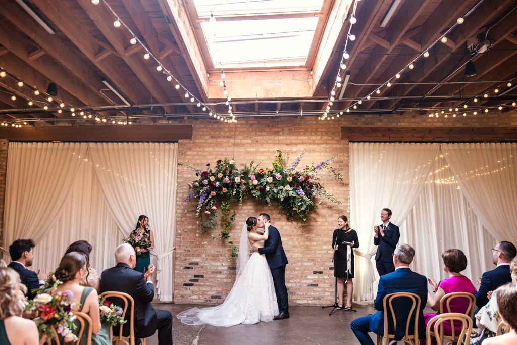 Bride and groom kiss during their wedding ceremony at The Arbory Chicago as guests watch and applaud