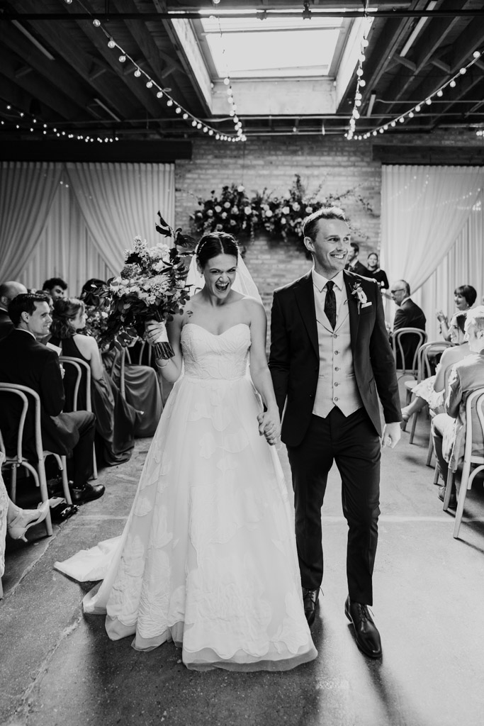 Black and white photo of happy newlyweds walking down the aisle smiling, surrounded by seated guests aduring their wedding ceremony at The Arbory Chicago