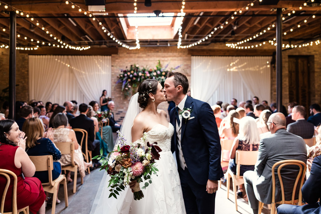 Bride and groom kiss while walking down the aisle during their wedding ceremony at The Arbory Chicago, with guests seated