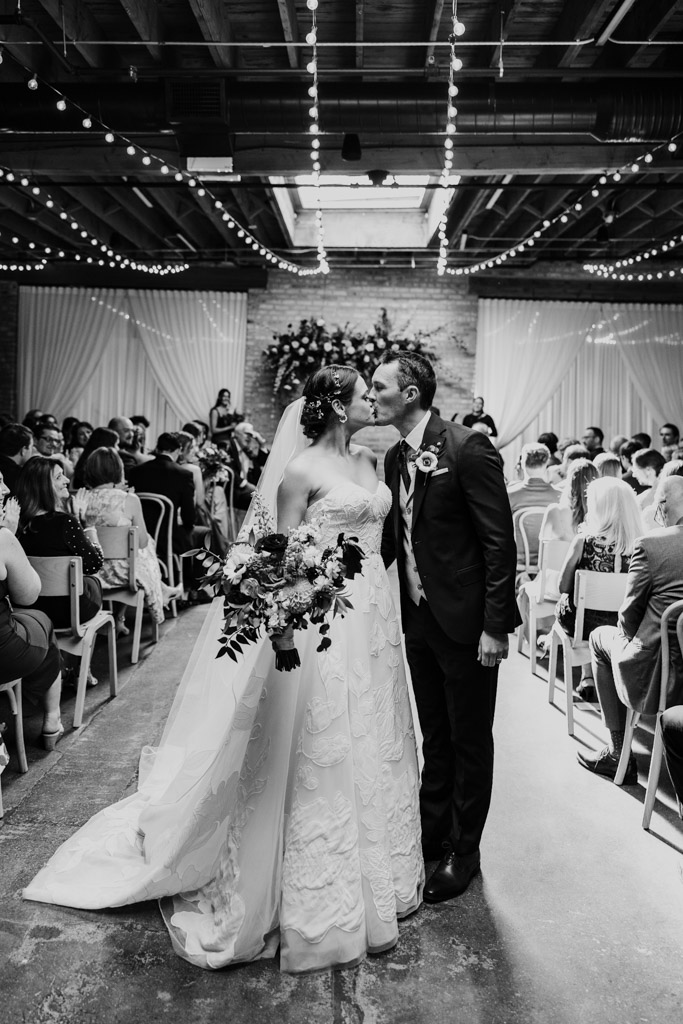 Black and white photo of bride and groom kissing at the end of the aisle, surrounded by guests during their wedding ceremony at The Arbory Chicago
