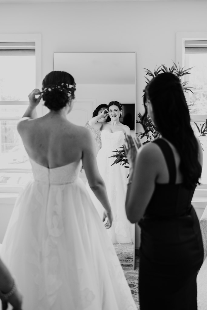 Black adn white photo of bride in a gown smiling in a mirror as her mother and sister help her get ready for her wedding celebration at The Arbory Chicago