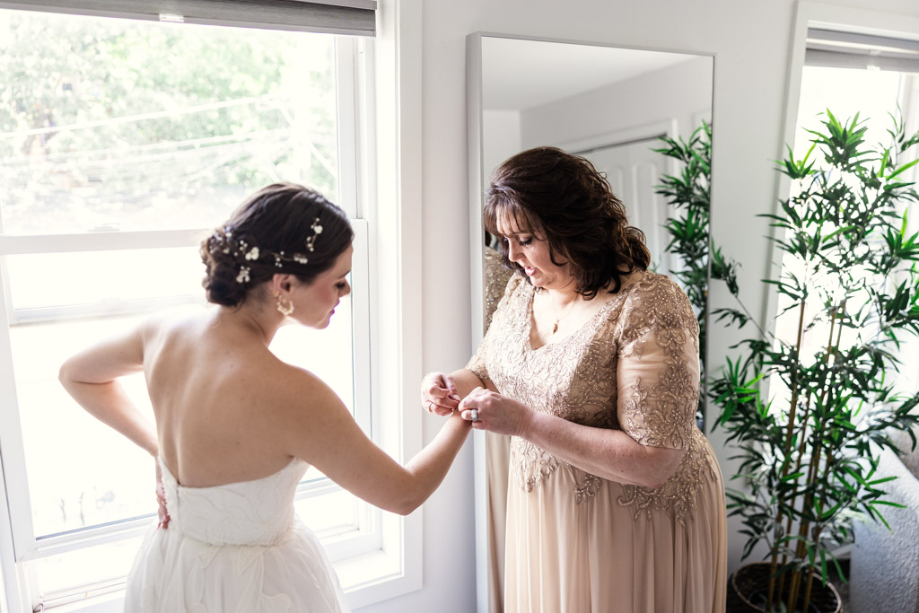 Bride's mother helps bride with her bracelet while they stand by a window in a bright room before wedding celebration at The Arbory Chicago