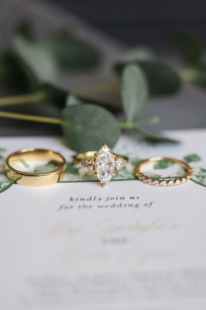 Detail photo of three gold rings, including a marquise diamond engagement ring, displayed on a wedding invitation with greenery