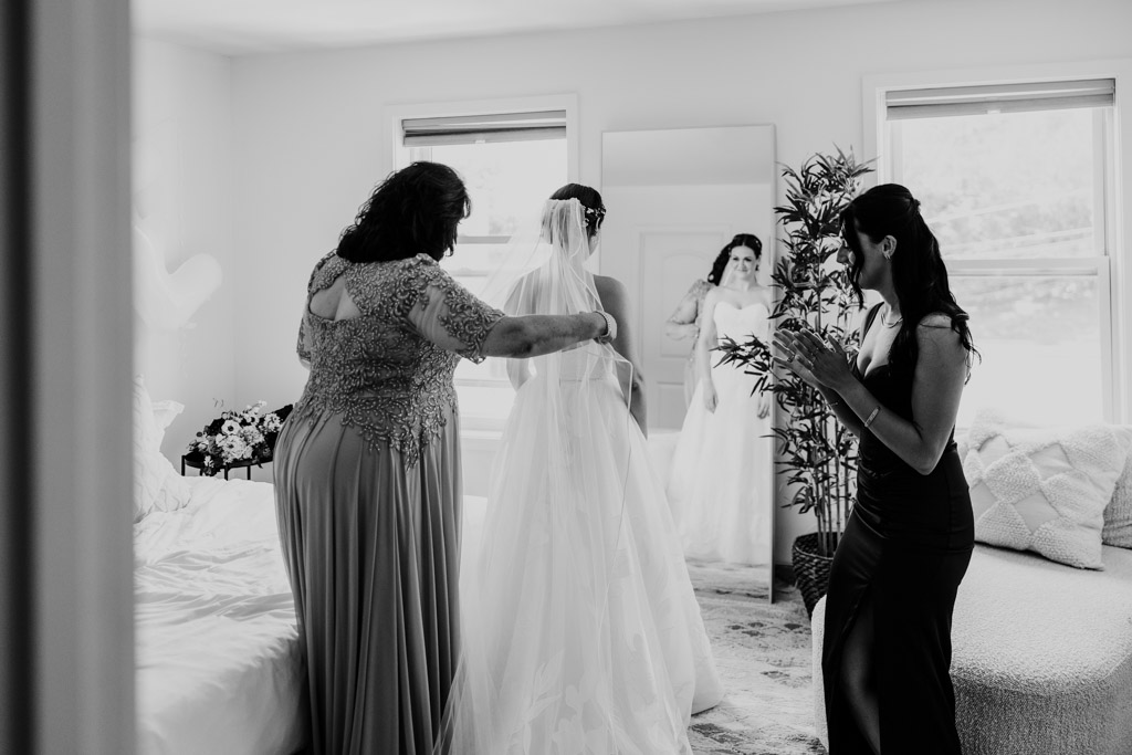 Black and white photo of bride getting ready with help from her mother and sister in a bright room