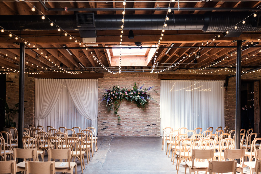 Wedding ceremony space at The Arbory Chicago with wooden chairs, string lights, draped curtains, and a floral arrangement on a brick wall