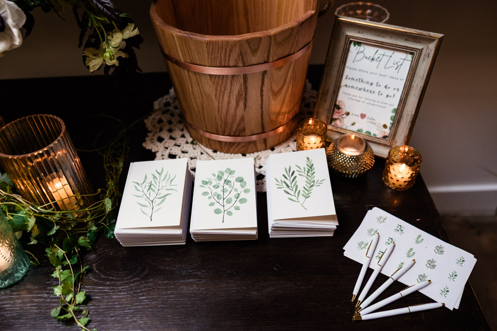 Wooden bucket, candles, plant-themed cards, and matching pens arranged on a dark table with a framed sign for wedding reception at The Arbory Chicago