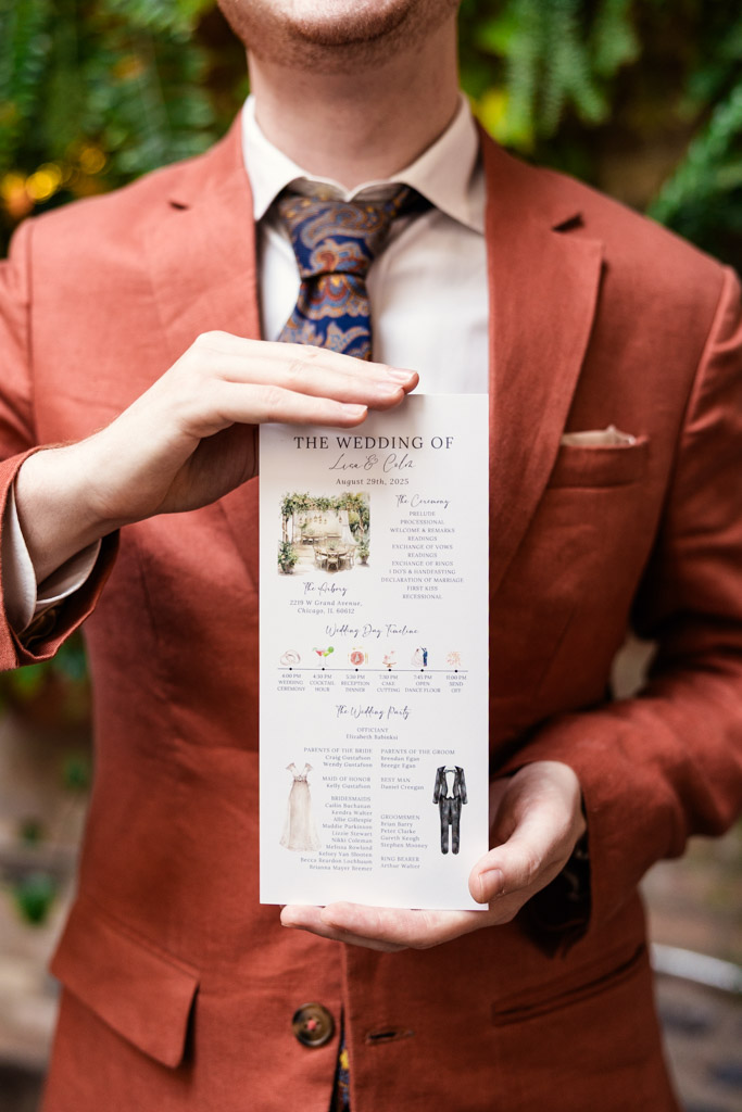 Guest in a rust-colored suit holding a wedding program with greenery in the background at The Arbory Chicago