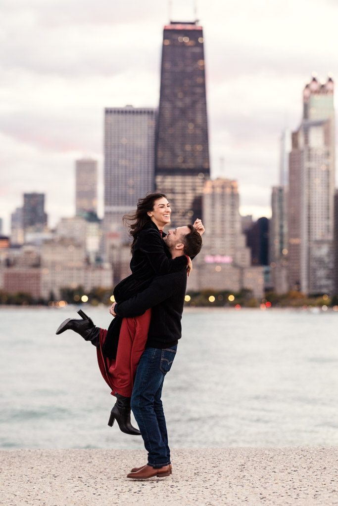 Man lifts his smiling partner by Lake Michigan with tall downtown Chicago buildings in the background during their fall engagement session