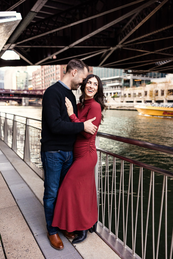 A couple embraces and smiles by a Chicago Riverwalk railing under a city bridge during their downtown Chicago fall engagement session