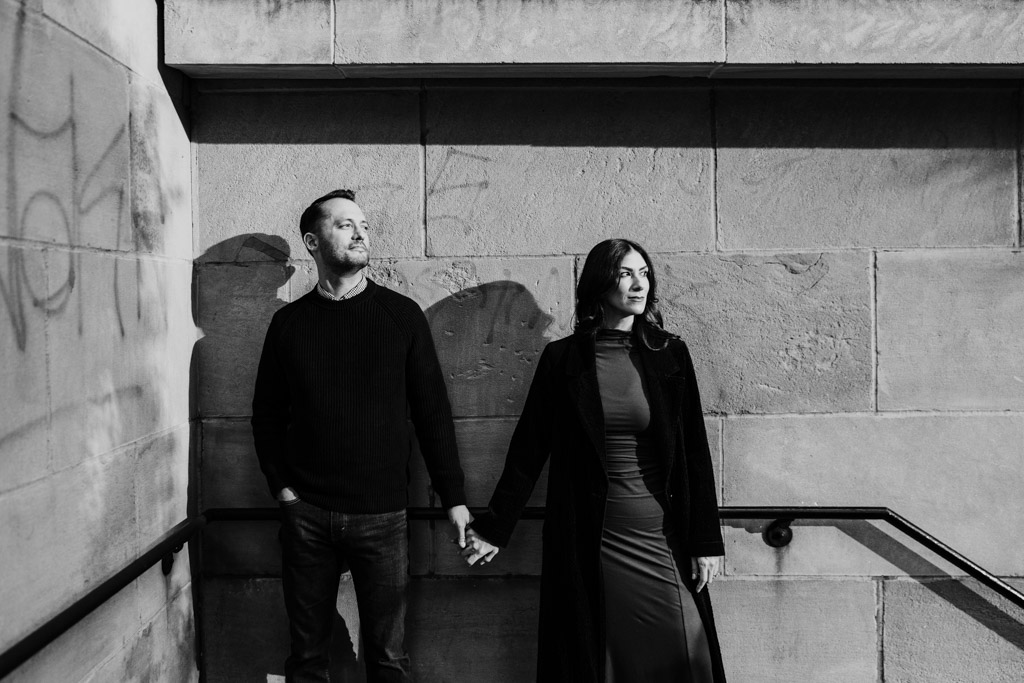 Black and white photo of couple holding hands stands against a sunlit stone wall during their downtown Chicago fall engagement