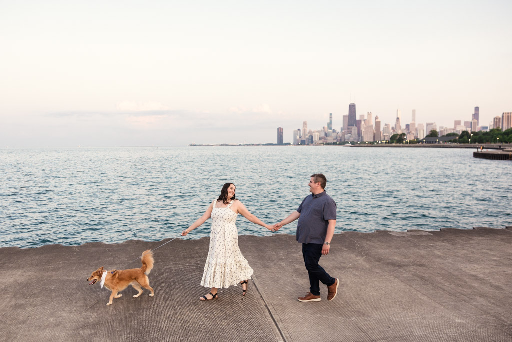 A couple holding hands and walking a dog by the water, with the Chicago skyline in the background, enjoying a perfect summer engagement.