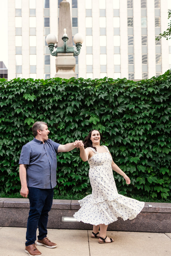 A man and woman dance together on a city sidewalk in front of a green ivy-covered wall, celebrating their Chicago summer engagement.