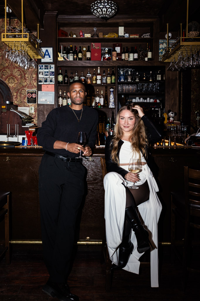 A man and a woman pose at the bar inside Emilio's Ballato during their New York City engagement session, with shelves of bottles behind them