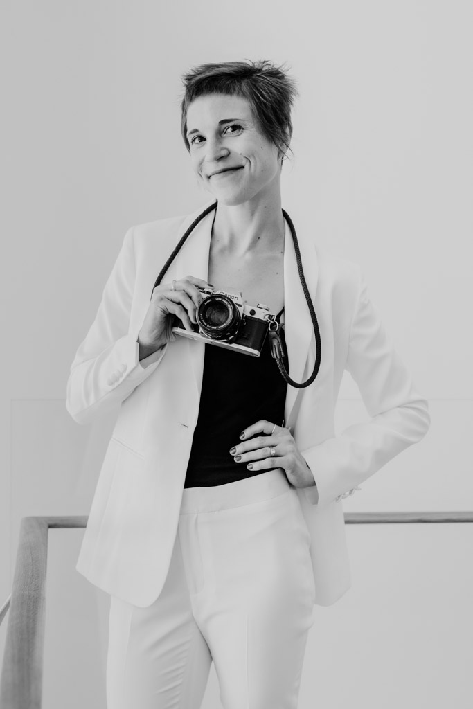 Black and white photo of a documentary wedding photographer in a white suit, holding a camera and smiling at the lens.