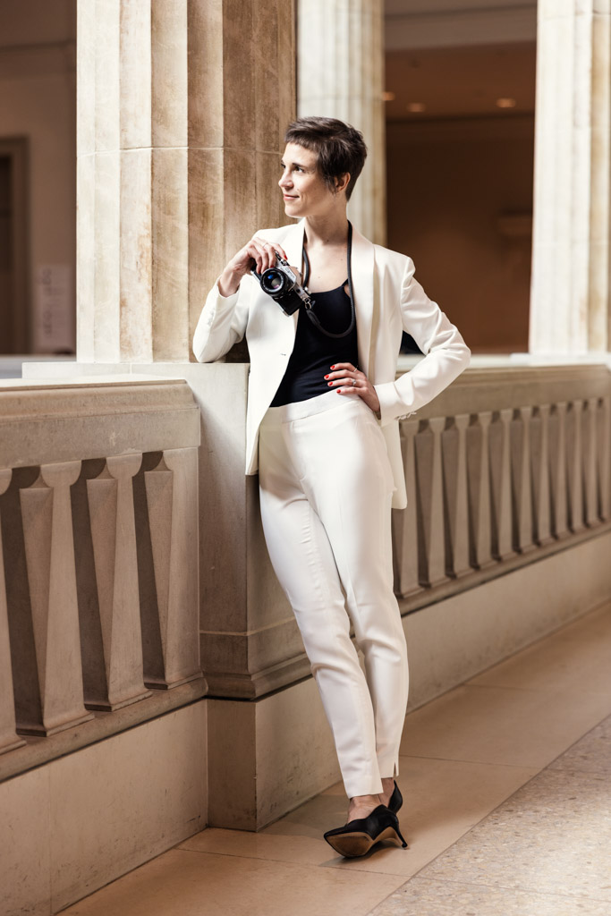 Emma Mullins in a white suit stands by a stone railing in the Art Institute of Chicago, holding a camera and looking to the side
