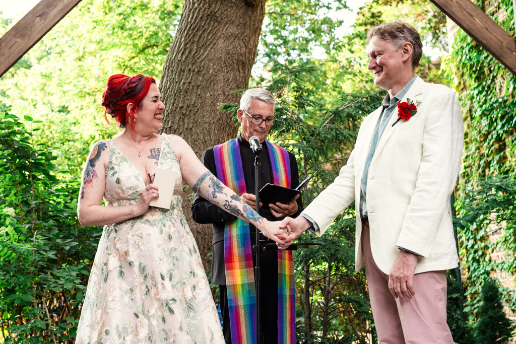 A couple holding hands and smiling during an outdoor wedding ceremony at Firehouse Chicago, with an officiant in a rainbow stole standing behind them