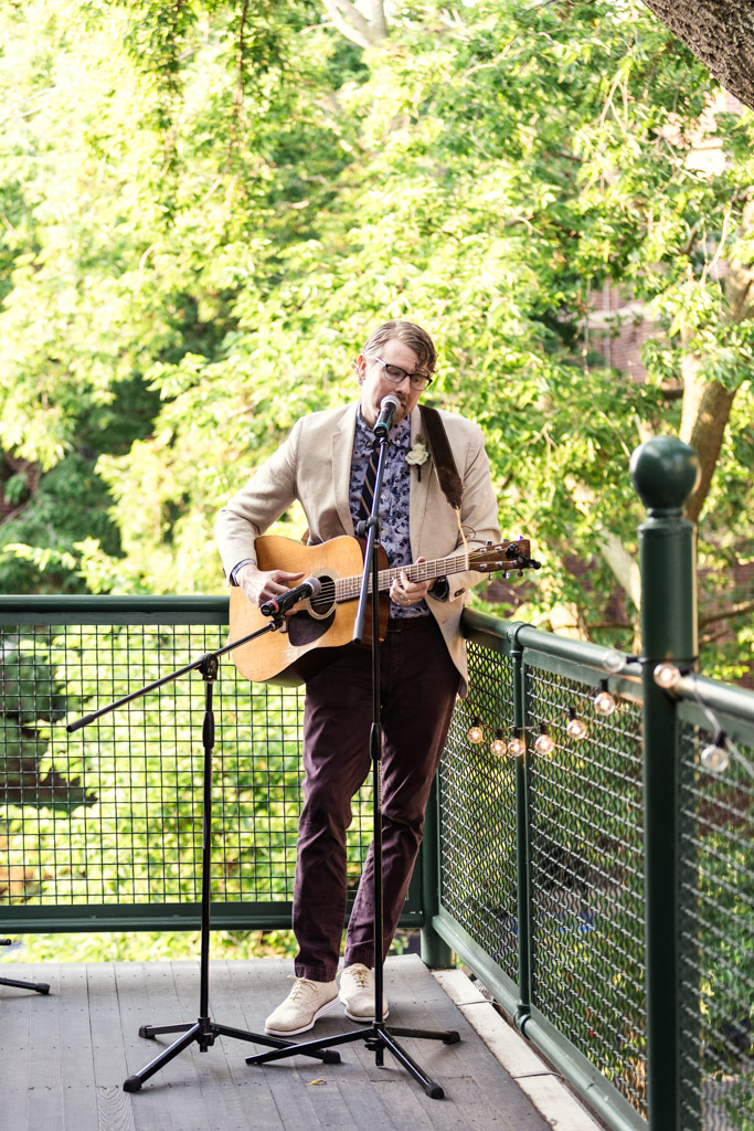 Man in a light blazer plays acoustic guitar and sings into a microphone on an outdoor deck at Firehouse Chicago