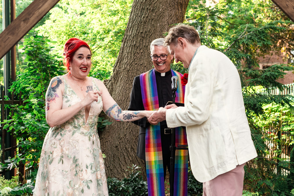A smiling couple exchanges rings at an outdoor Firehouse Chicago wedding, with an officiant in a rainbow stole standing behind them