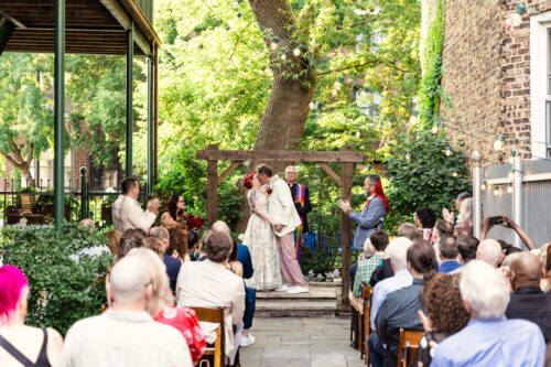 A couple kisses at their outdoor Firehouse Chicago wedding ceremony as guests seated on both sides watch and applaud.