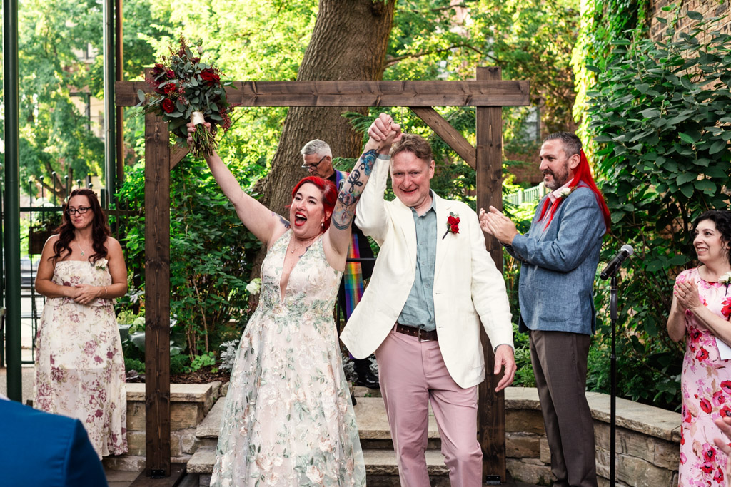 Happy newlywed couple celebrates their wedding at Firehouse Chicago, holding hands up, surrounded by smiling guests in a lush garden