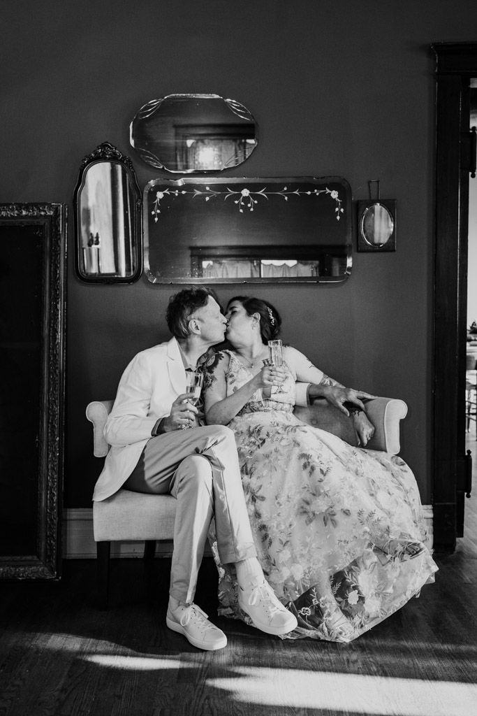 Black and white photo of bride and groom dressed formally kissing on a loveseat at Firehouse Chicago, holding drinks and surrounded by vintage mirrors on the wall